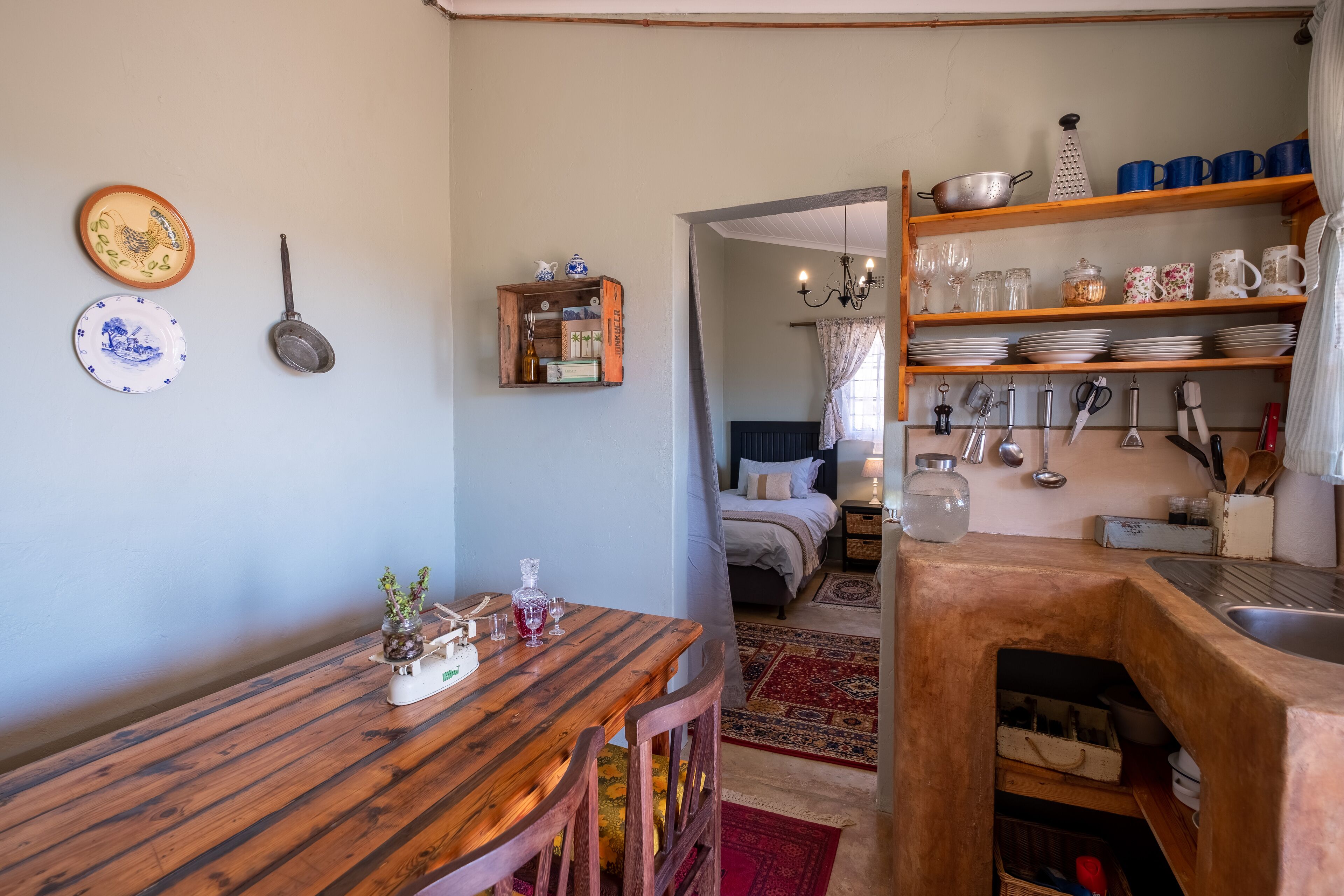 A cozy kitchen with wooden shelves, a dining table, and a bed in the background.