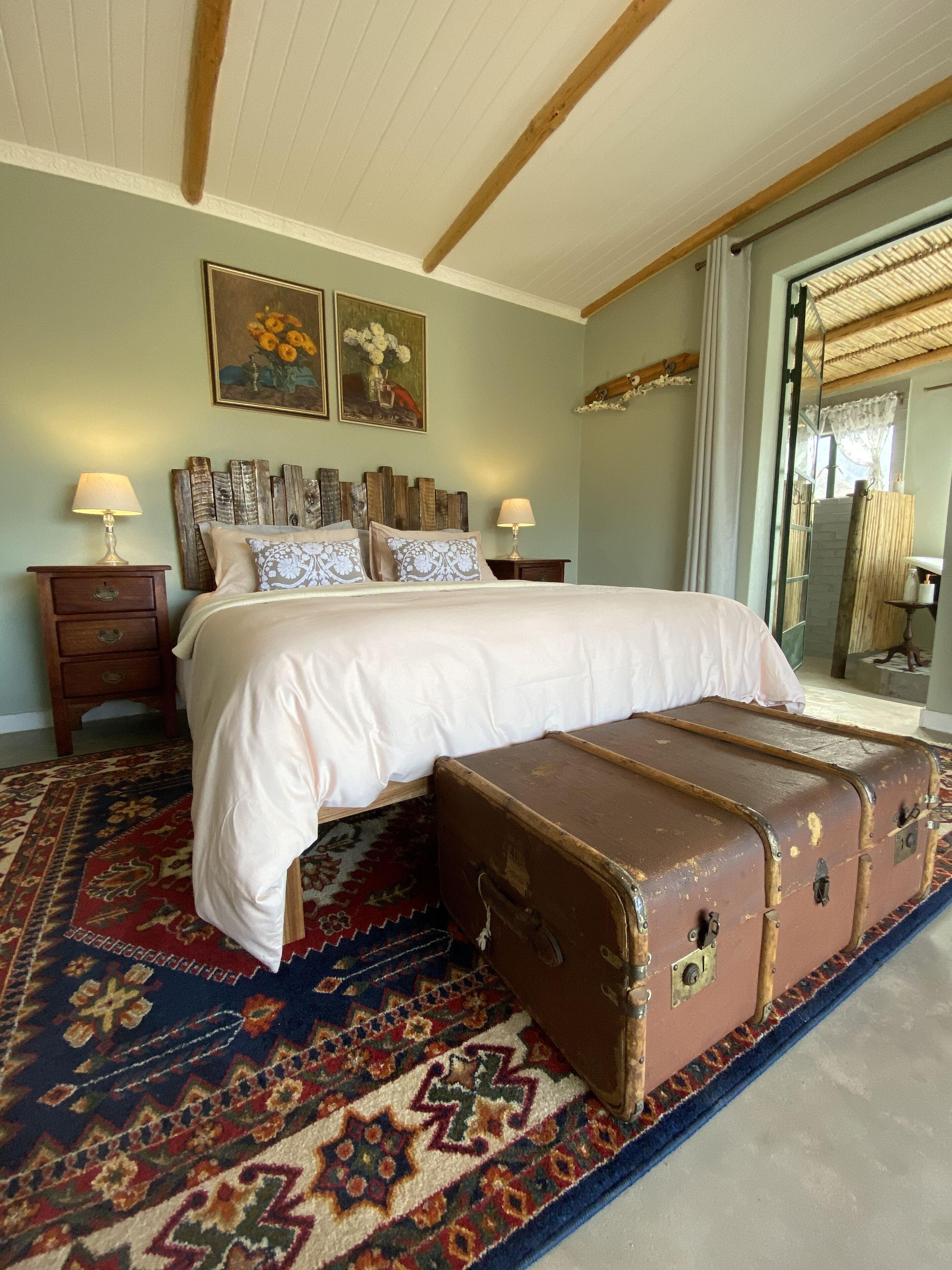 A bedroom with a large bed, a wooden headboard, two bedside lamps, a patterned rug, and a vintage suitcase.