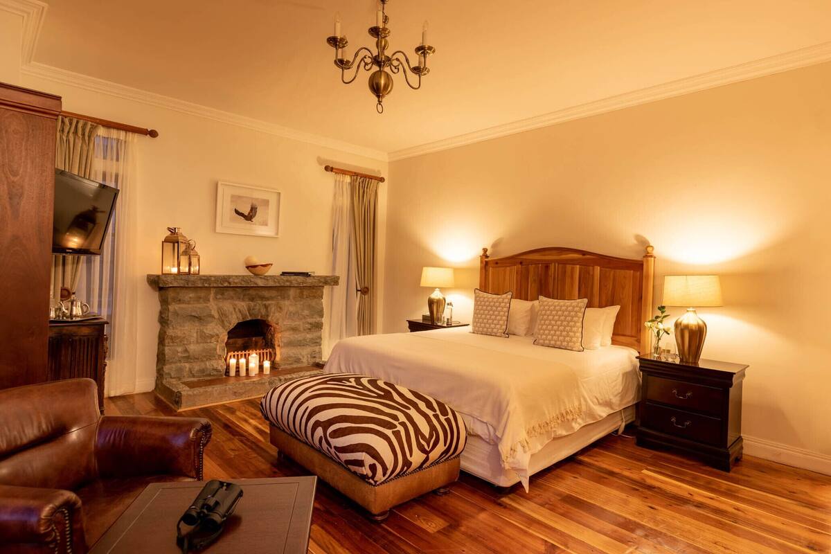 A cozy bedroom with a stone fireplace, a large bed with two bedside tables, a leather armchair, and a wooden floor.