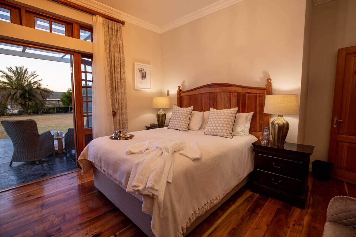 A bedroom with a large bed, two bedside tables with lamps, a wooden headboard, and a view through a glass door to the outside.