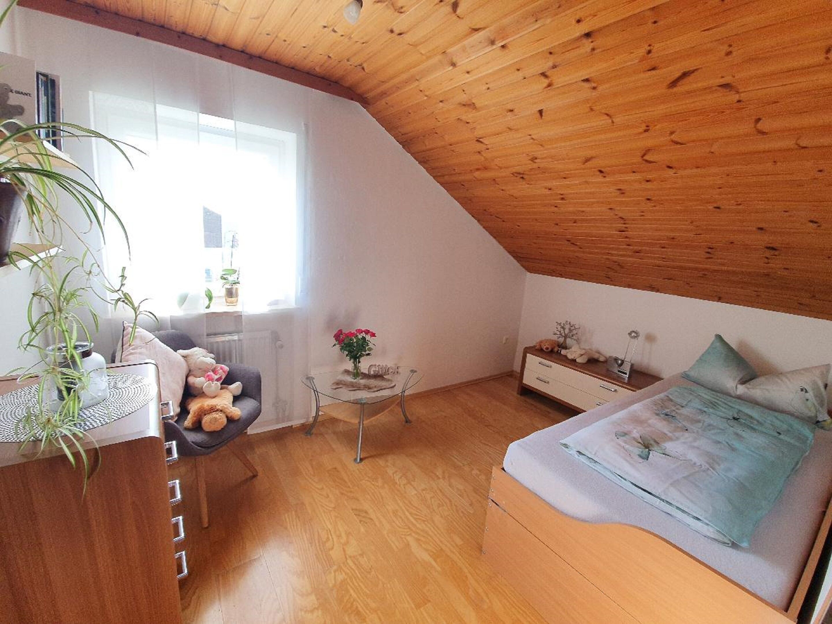 A cozy attic bedroom with a sloped ceiling, a bed, a small table with a vase, and a dresser.