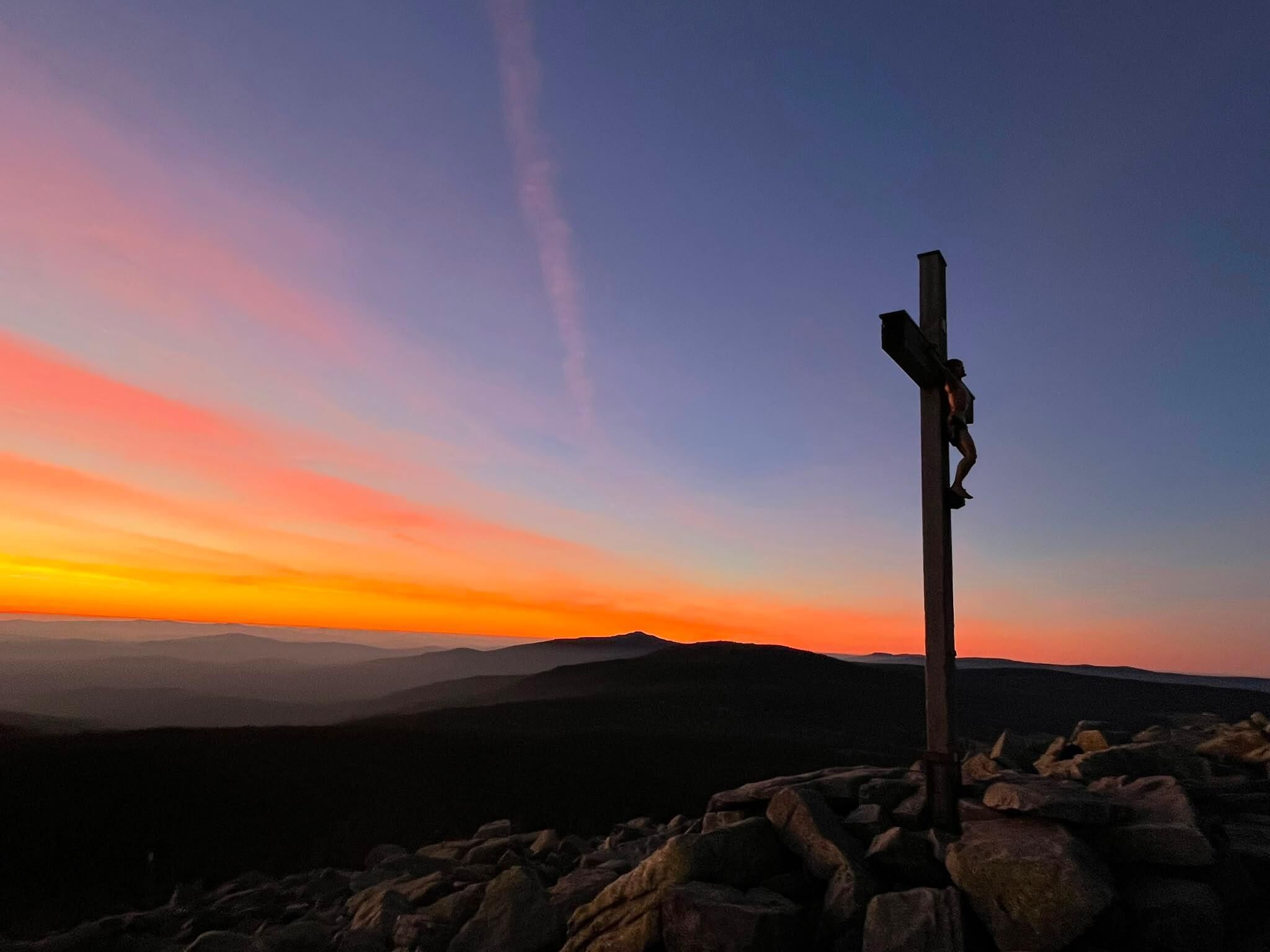 Une croix au sommet d’une montagne, avec un coucher de soleil en arrière-plan.