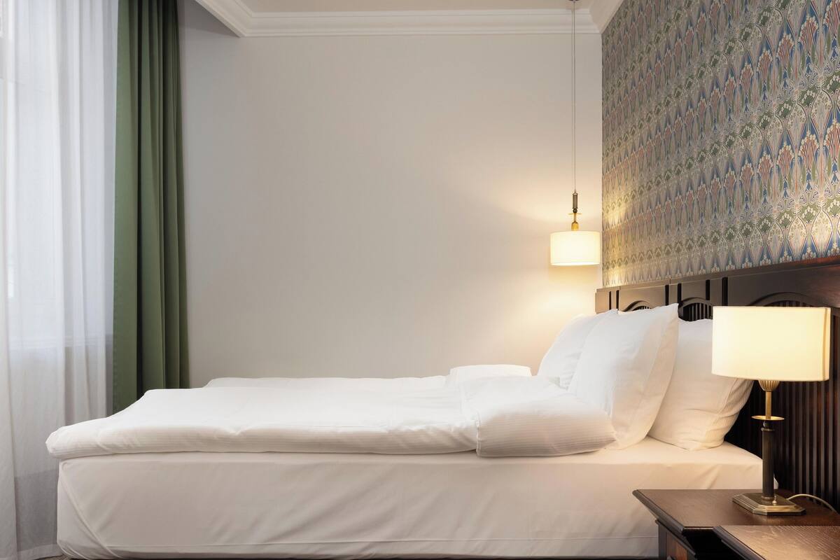 Una cama bien tendida con sábanas blancas, una mesita de noche con una lámpara y un papel tapiz estampado en la pared.