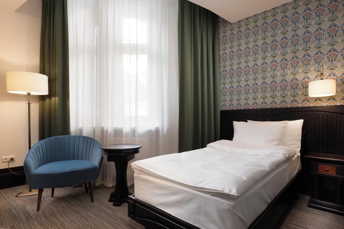 Una habitación de hotel con una cama, una silla, una mesita de noche, una lámpara y un ventanal con cortinas.