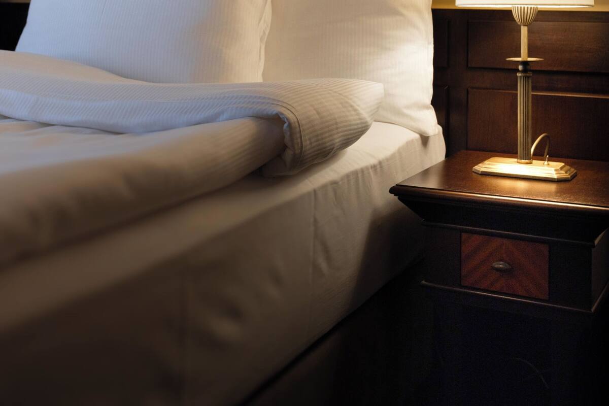 Una cama bien tendida con sábanas y almohadones blancos, una mesita de noche con lámpara y un mueble auxiliar de madera.