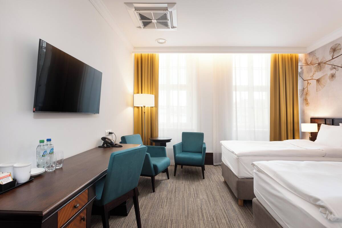 Habitación de hotel con televisión de pantalla plana, dos camas individuales, un escritorio con sillas, una mesita y una lámpara.
