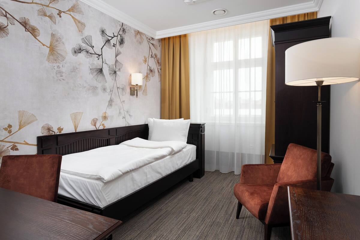 Una habitación de hotel con cama, escritorio, silla, lámpara y ventana con cortinas.