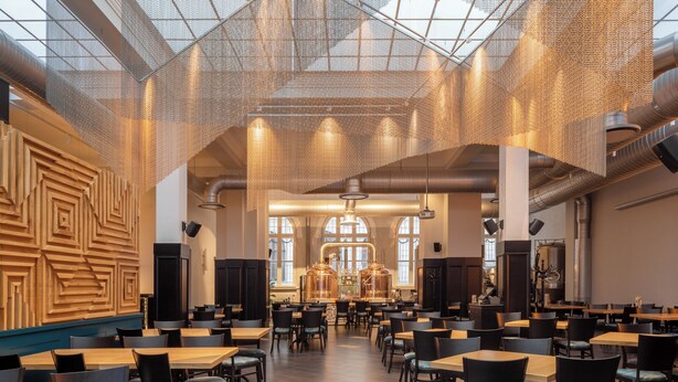 A spacious indoor area with high ceilings, a patterned wall, and multiple tables and chairs arranged for dining or seating. - Brovaria (Poznan)