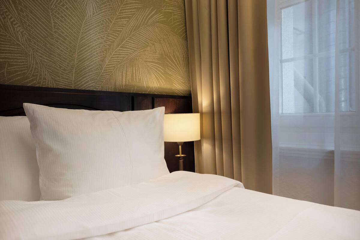 Una cama bien hecha con sábanas blancas, una lámpara de noche y una ventana con cortinas translúcidas.