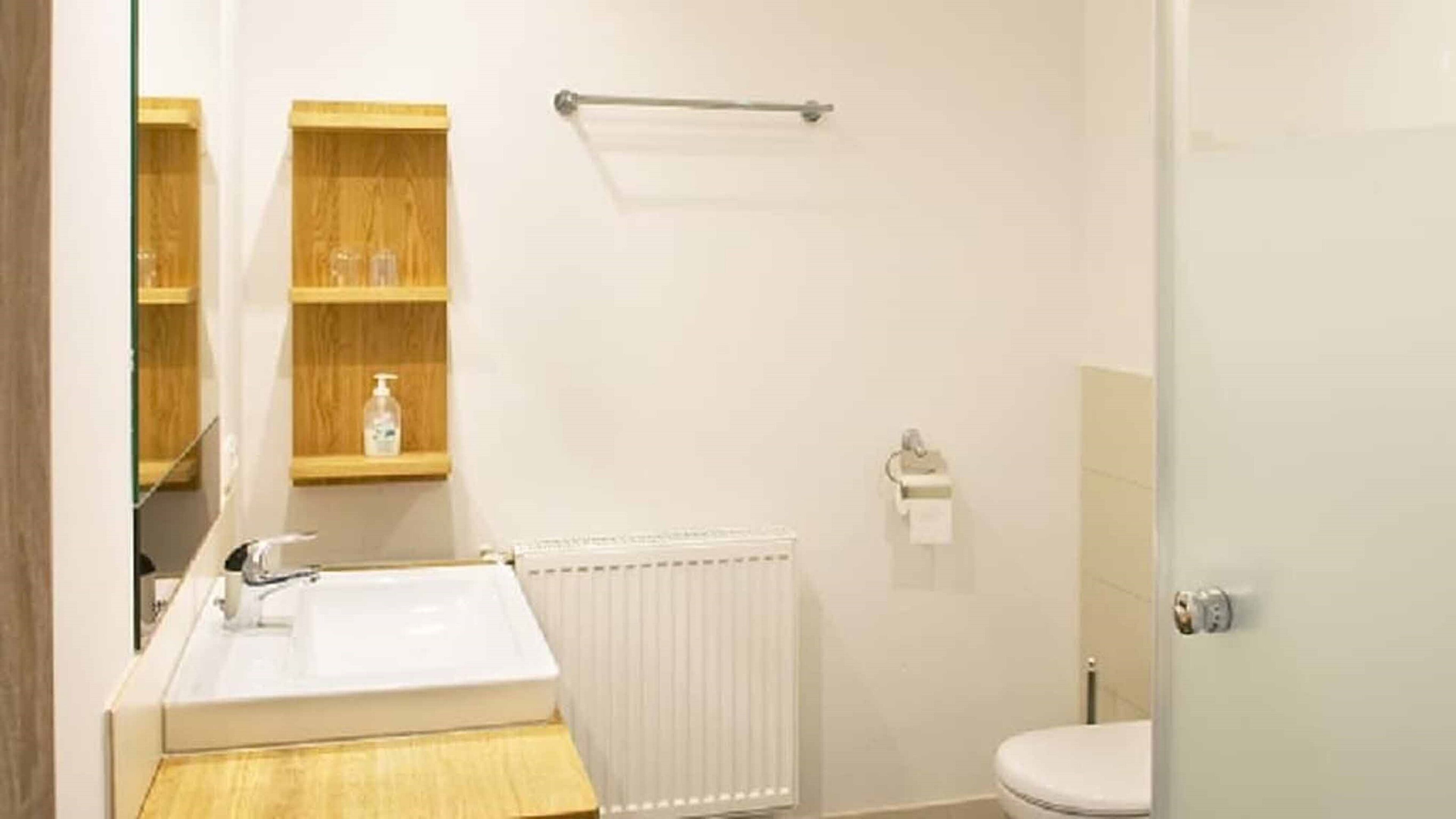 Un baño con lavamanos blanco, estantes de madera, un perchero para toallas y un inodoro.