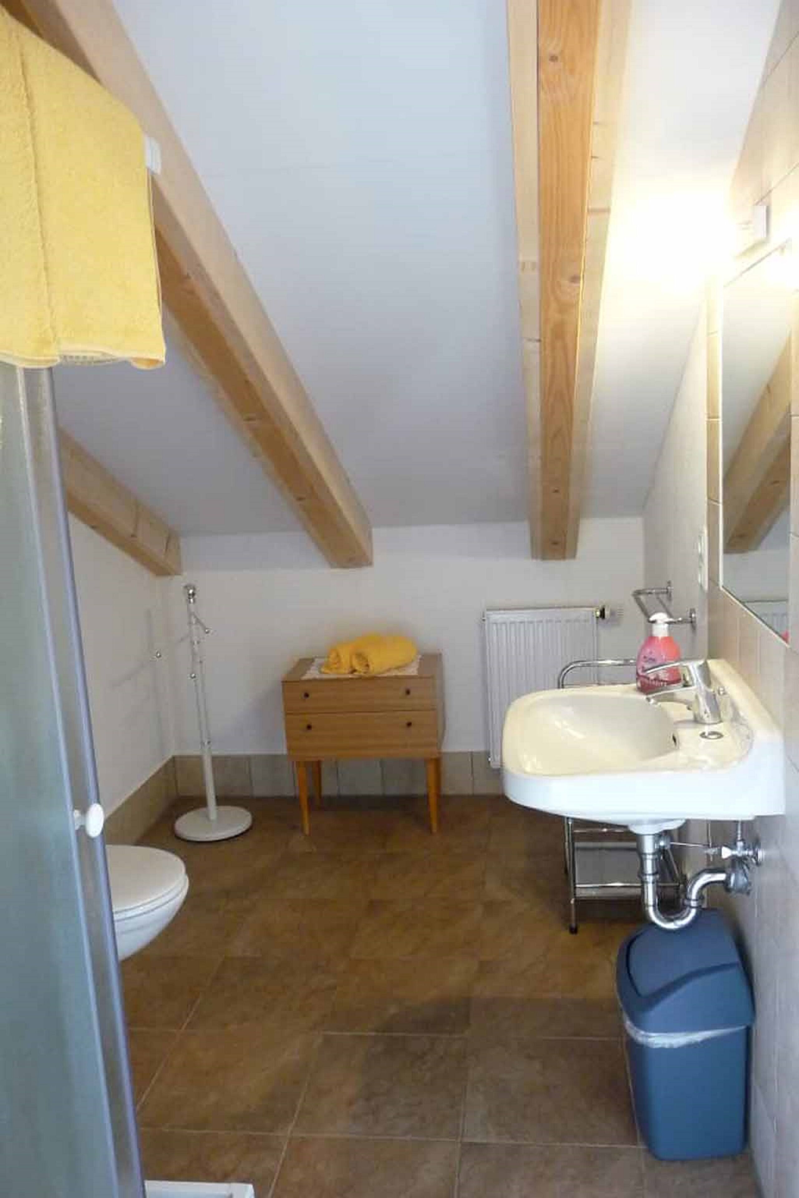 A small bathroom with a white sink, wooden beams, a yellow towel, and a blue trash bin.