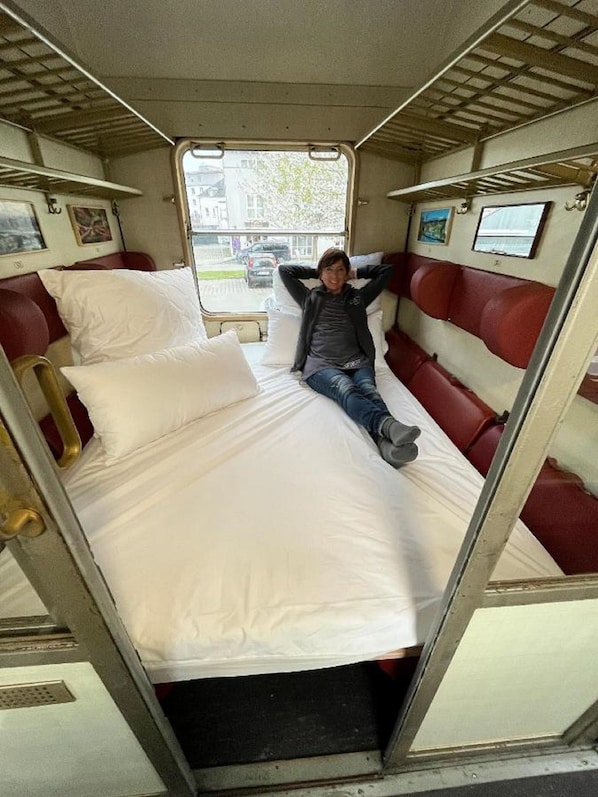 A narrow, enclosed sleeping area with a single bed, red and white headboards, and a window at the far end. - Übernachtung im nostalgischen Bahnwaggon (Bogen)
