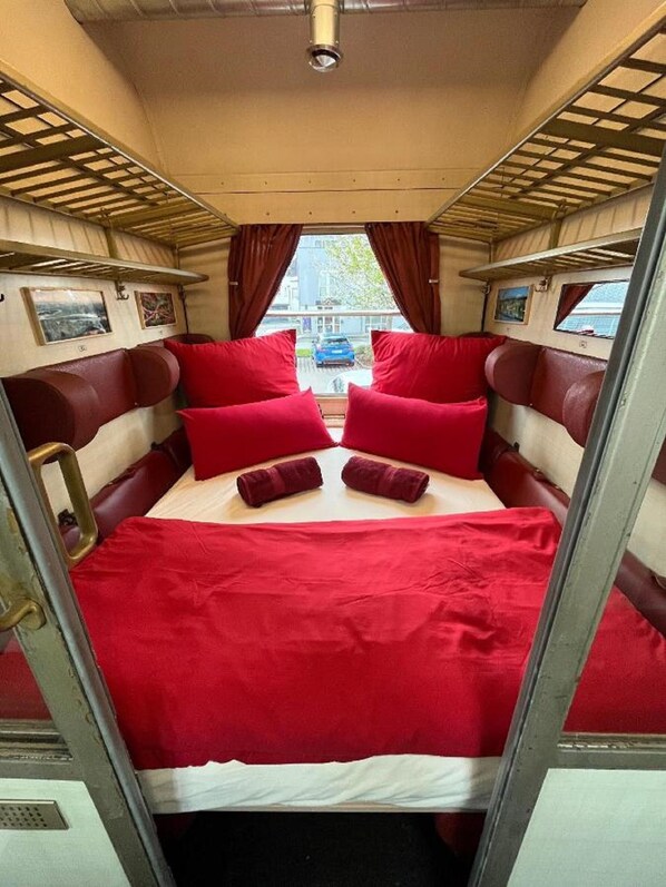 A narrow train compartment with a bed, red bedding, and curtains. - Übernachtung im nostalgischen Bahnwaggon (Bogen)