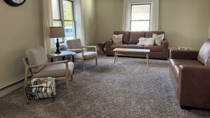 A living room with a brown leather sofa, a wooden coffee table, and a side table with a lamp. There is a bookshelf with books and a wall clock. - Heart of Houghton! Trails, Shopping & More! (Dodgeville)
