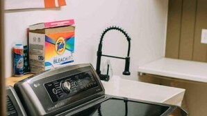 A laundry area with a front-loading washing machine and dryer, a wall-mounted faucet, and a shelf with cleaning supplies. - Heart of Houghton! Trails, Shopping & More! (Dodgeville)