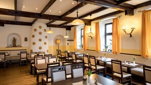 En restaurant med træborde og stole, hvælvet loft med synlige bjælker samt vægmonterede lamper.
