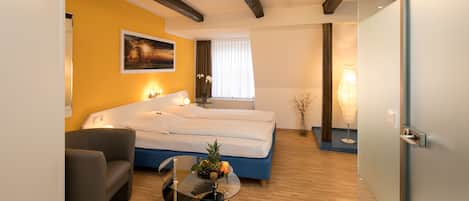 A modern hotel room with a bed, a sofa, a glass coffee table with fruit, and a painting on the wall.