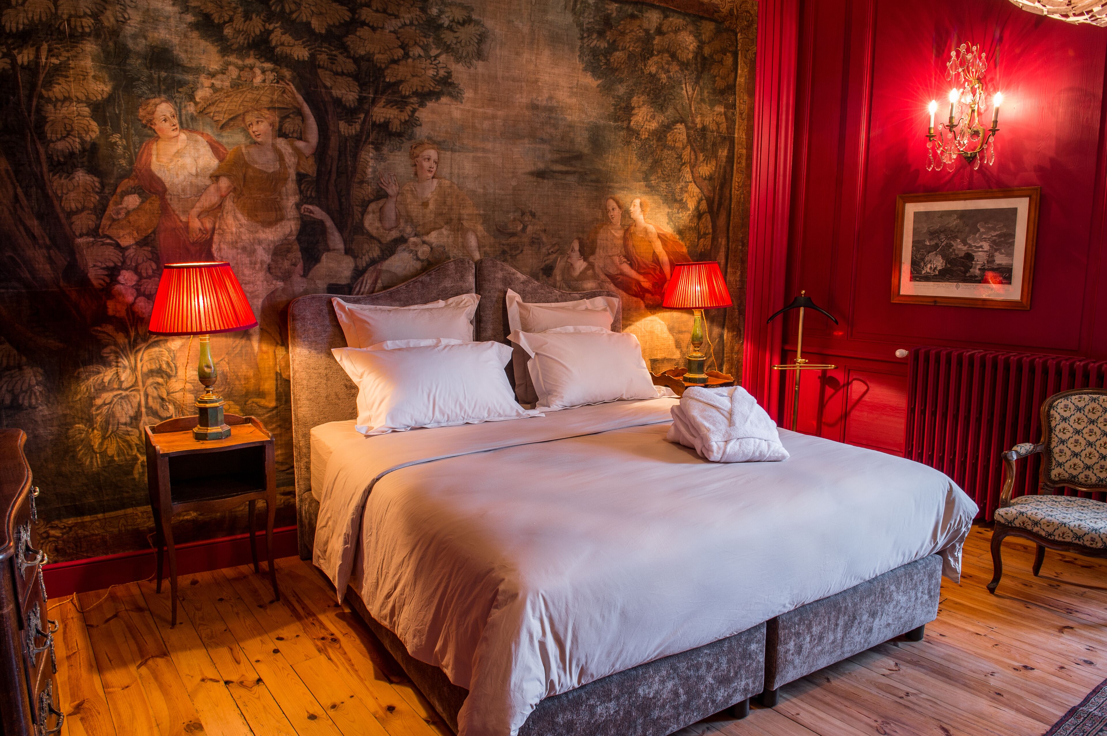 A large bed with white linens, two bedside tables with lamps, a red wall with a mural, and a chair.