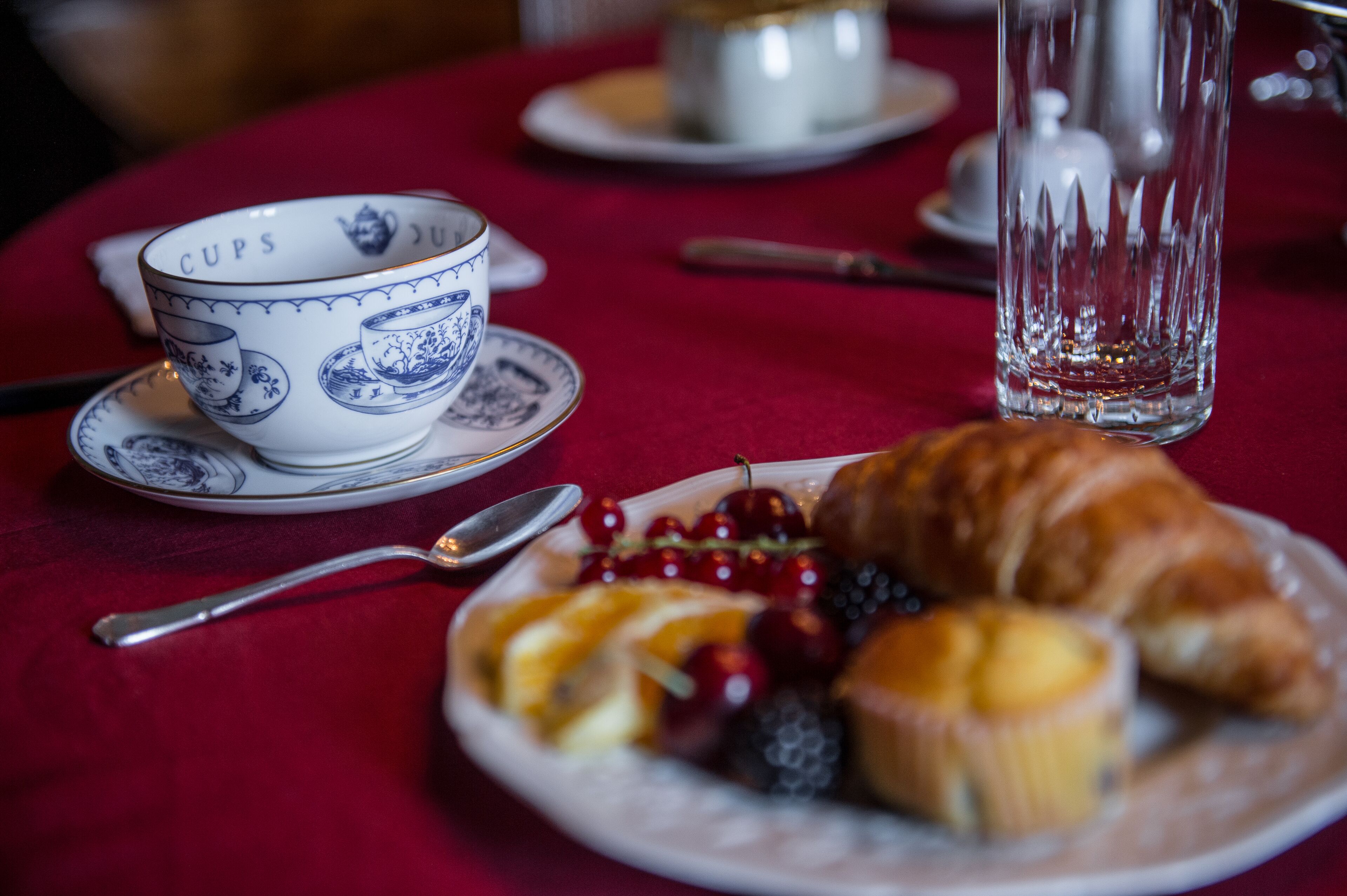 Une table recouverte d’une nappe rouge, une assiette de fruits et un croissant, une tasse à motifs bleus et un verre transparent.