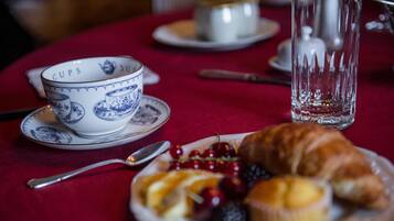 Una mesa con mantel rojo, un plato de fruta y un croissant, una taza con diseño azul y un vaso transparente.