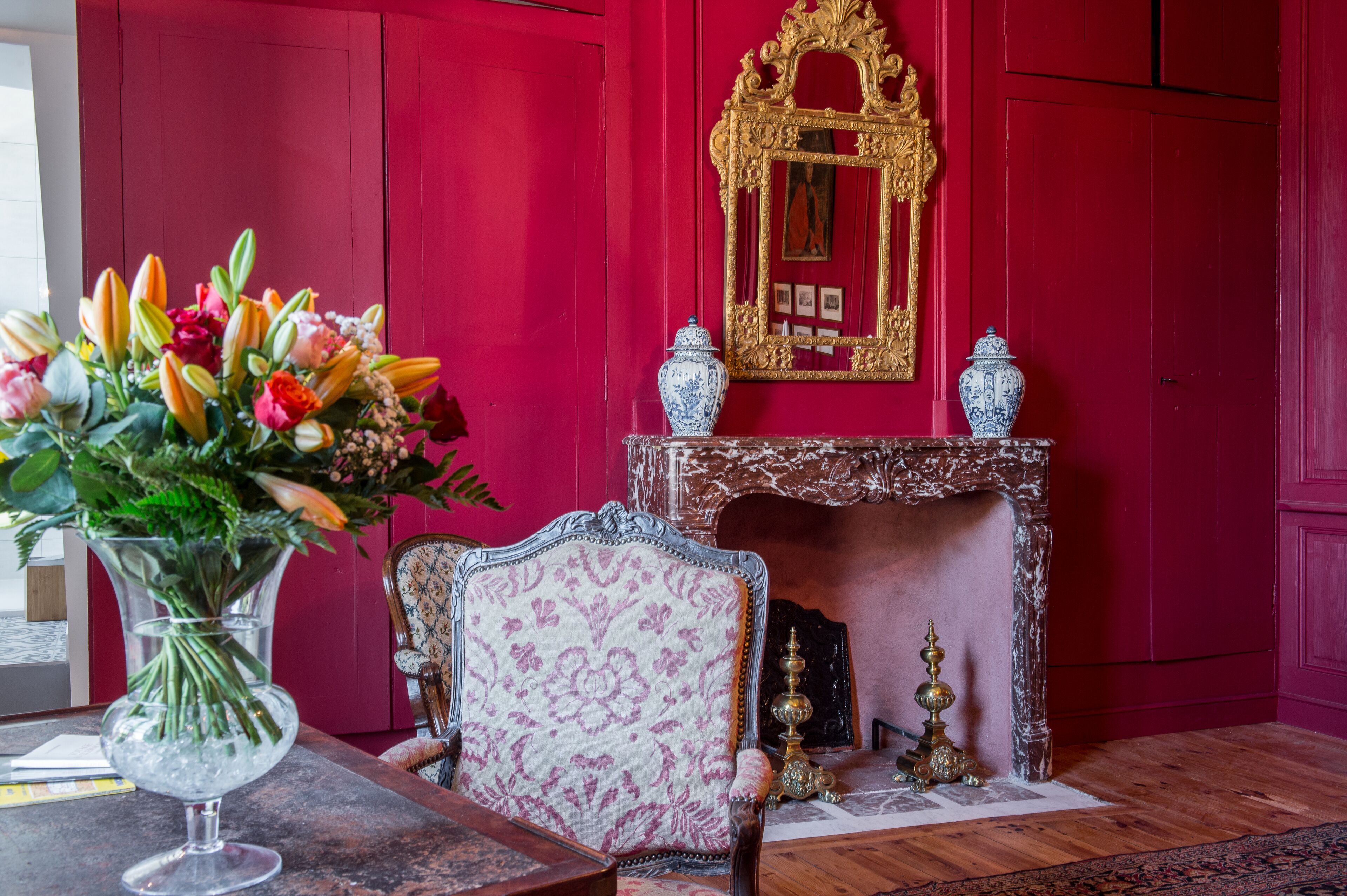 Une pièce avec un mur rouge, une cheminée, un miroir et un arrangement floral sur une table.