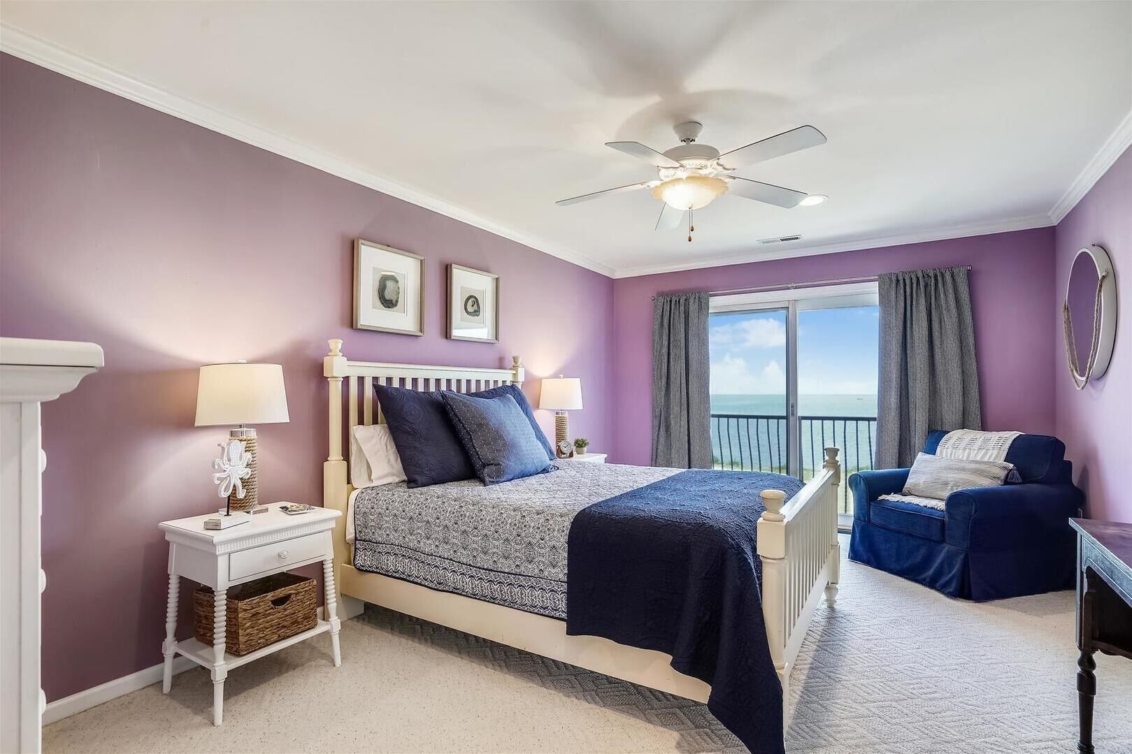 A bedroom with a bed, nightstands, a dresser, a chair, a ceiling fan, and a view of the sea through a balcony.