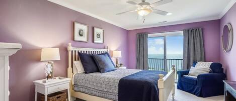 A bedroom with a bed, nightstands, a dresser, a chair, a ceiling fan, and a view of the sea through a balcony.