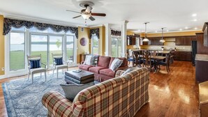 A spacious living room with a large sofa, a coffee table, and a view of a grassy area through large windows. - Minutes from Downtown Cambridge | Private Dock | Pool | Kayaks (Cambridge)