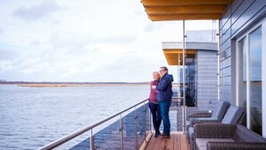 Een stel staat op een dek met uitzicht op een kalme waterpartij en veraf gelegen land.