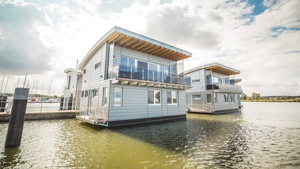 Een drijvend woonschip met een balkon en grote ramen, afgemeerd in een jachthaven, met boten op de achtergrond.