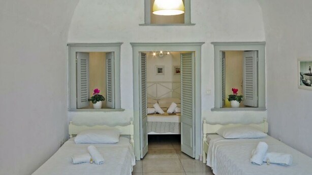 A room with two beds, a small bathroom, and a window with shutters. - Nefeli Traditional Cave Houses (Santorini)