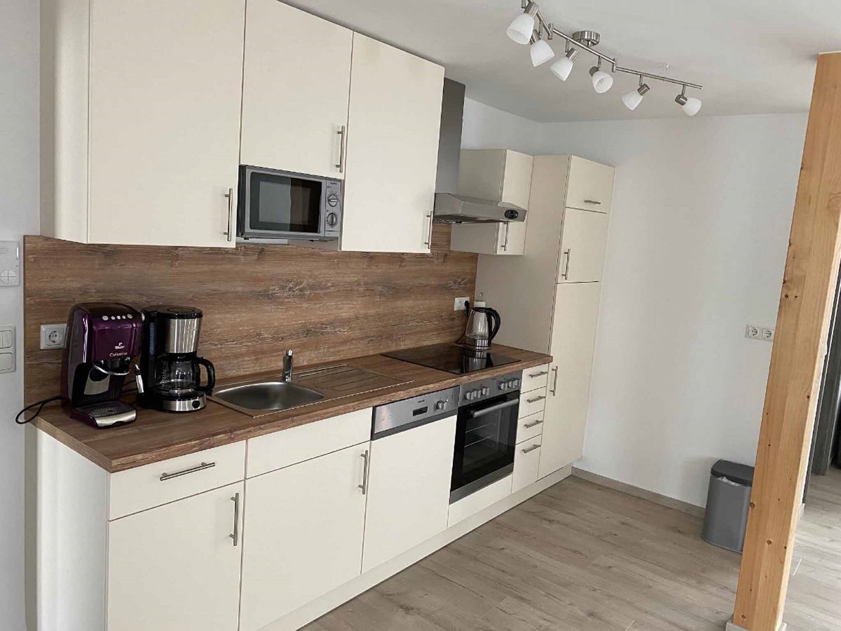 A compact kitchen with white cabinets, a wooden countertop, a built-in microwave, a coffee maker, a kettle, and a dishwasher.