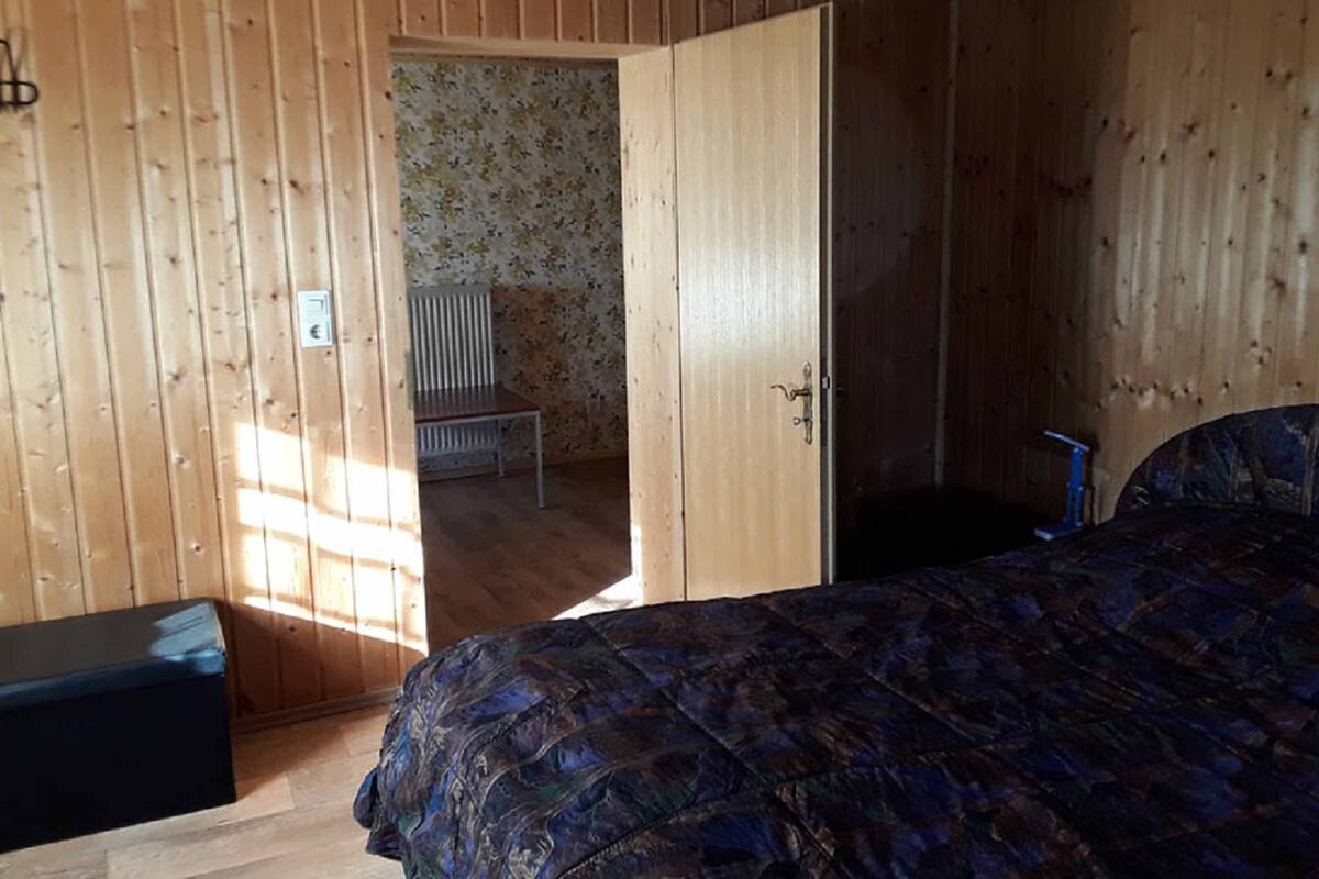 Une pièce en bois avec un lit, un banc et une porte donnant sur une autre pièce dont le mur est décoré de motifs floraux.