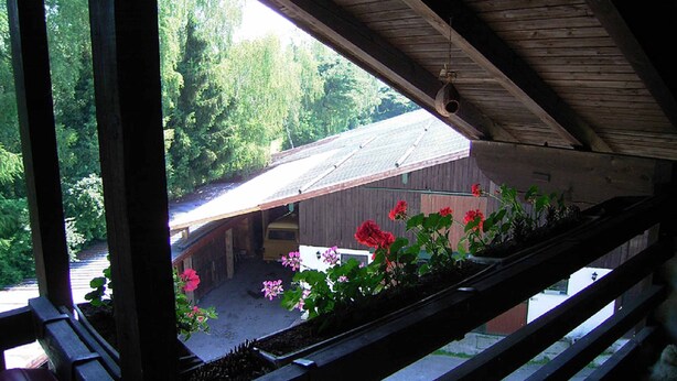 A view from a window with wooden beams and a balcony, overlooking a building with a sloped roof and a fenced yard with red flowers. - Naturfreundepension Hagenberger für Erwachsene (Witzmannsberg)