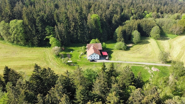 A house with a sloped roof, surrounded by a forested area and open fields.