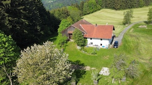A rural house with a red roof, surrounded by a green landscape and a gravel driveway.