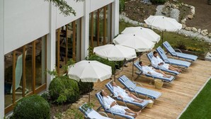 A deck with sun loungers and umbrellas, surrounded by a garden and a building with large windows. - Bio Thermalhotel Falkenhof (Bad Fuessing)