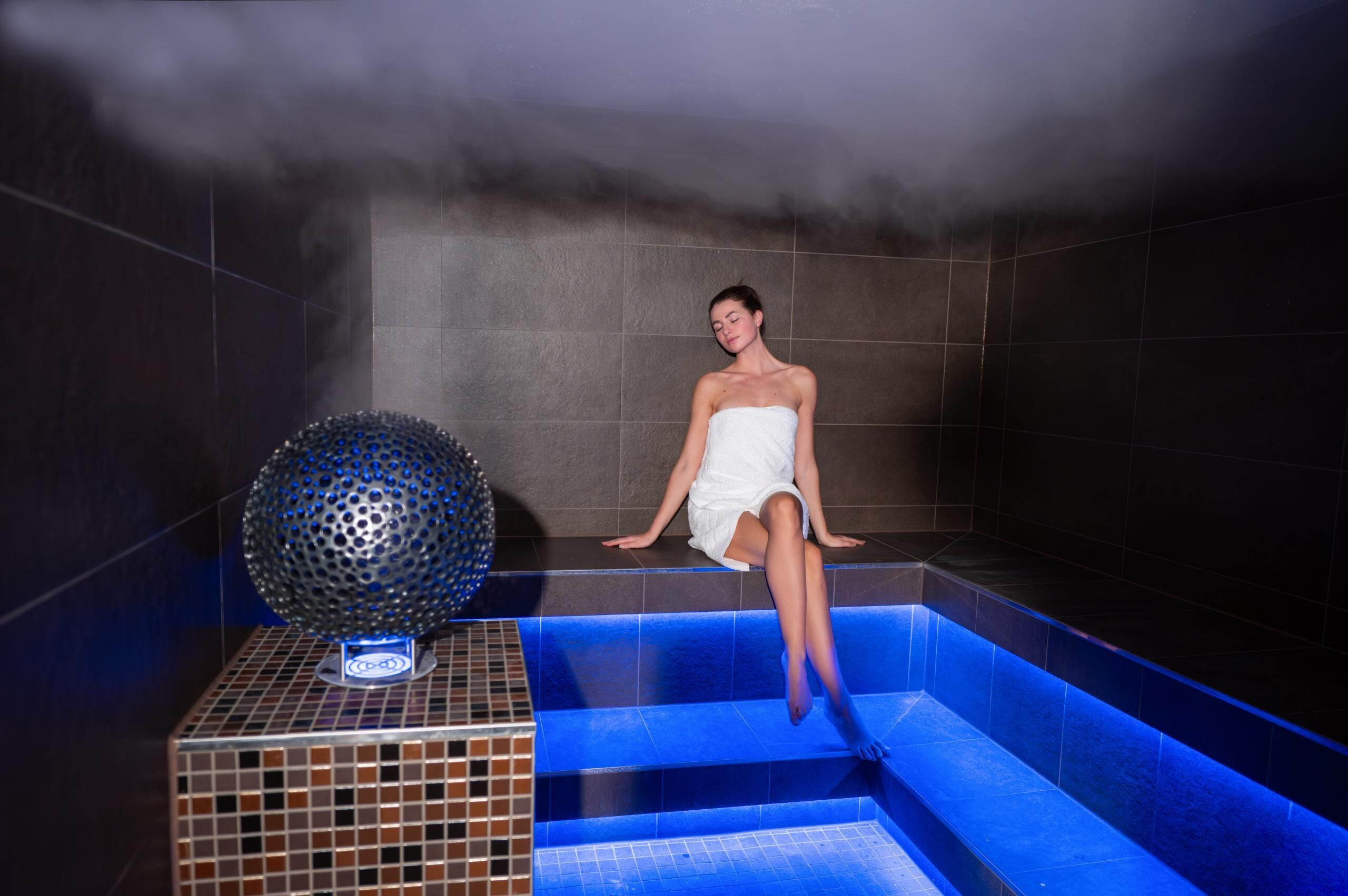Una persona sentada en una tina de hidromasaje con una fuente iluminada en azul y una esfera decorativa.