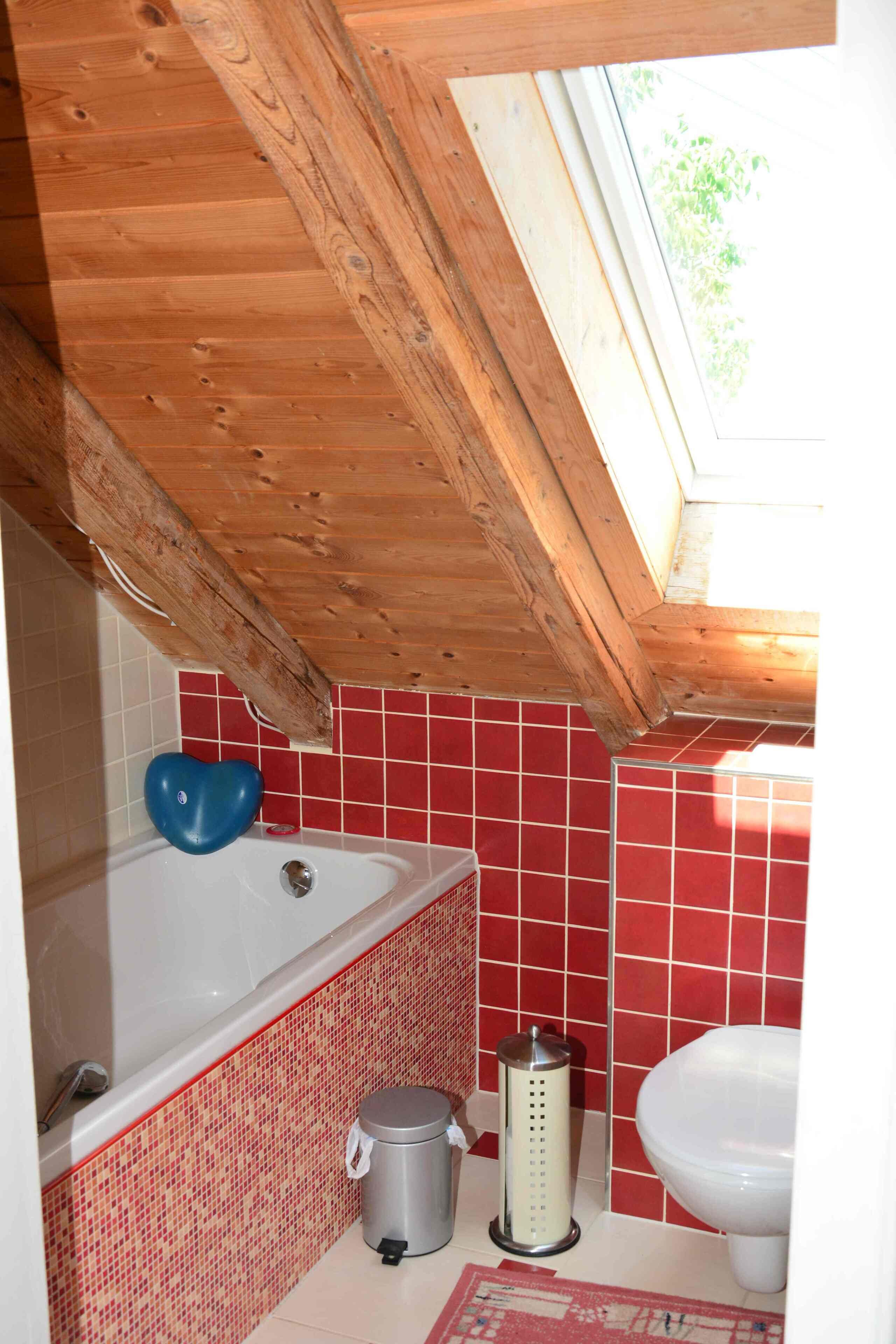 A bathroom with a wooden ceiling, red tiled walls, a bathtub, a toilet, and a trash bin.