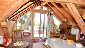 Una habitación cálida en el ático con vigas de madera, una mesa de comedor con fruta, un sofá y un ventanal corredero al exterior.