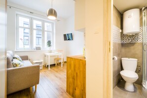 A modern living room with a sofa, a small table, and a TV. There is a wooden cabinet and a bathroom with a toilet and shower visible. - Solna Atelier (Kraków)