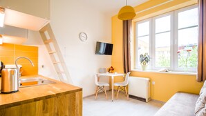 Cocina moderna con encimera de madera, hervidor, y un lavabo. Hay un comedor con una mesa y sillas, un televisor en la pared, y una ventana con vistas al exterior.