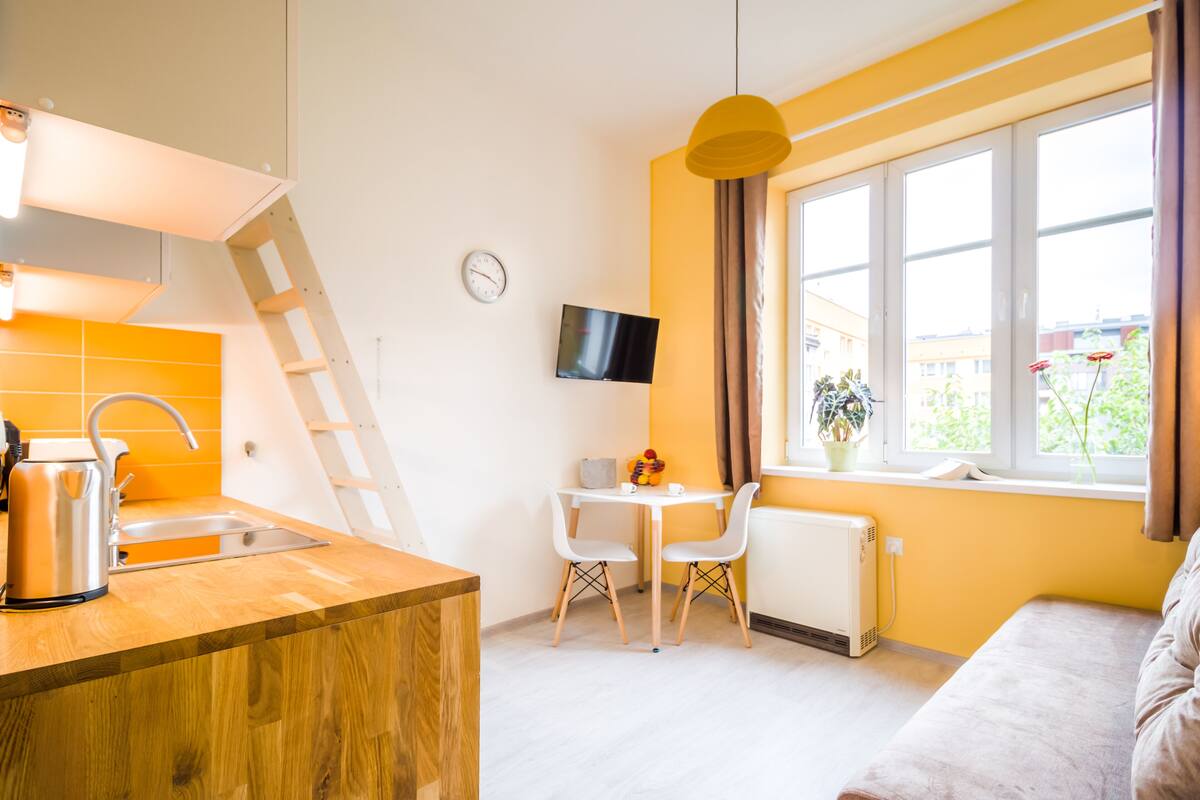 Cocina moderna con encimera de madera, hervidor, y un lavabo. Hay un comedor con una mesa y sillas, un televisor en la pared, y una ventana con vistas al exterior.
