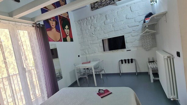 A compact hotel room with a white brick wall, a small window, a table with chairs, a radiator, and a curtain. - Old renovated studio apartment in Town Hall Square with works on display (Agrigento)