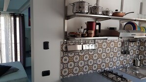 A kitchen with a gas stove, a sink, and various kitchen utensils. There are shelves above the counter holding pots and pans. - Old renovated studio apartment in Town Hall Square with works on display (Agrigento)
