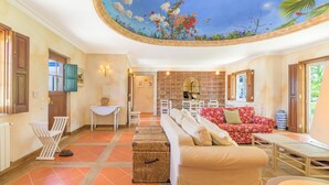 A spacious living area with a brick fireplace, a red floral sofa, and a ceiling mural. - Country House - Quinta das Tílias (Santiago do Cacém)