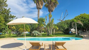 A poolside area with lounge chairs and a large umbrella, surrounded by palm trees and a clear blue sky. - Country House - Quinta das Tílias (Santiago do Cacém)