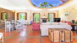 A spacious living area with a fireplace, a dining table with chairs, and a large window offering a view of greenery. - Country House - Quinta das Tílias (Santiago do Cacém)