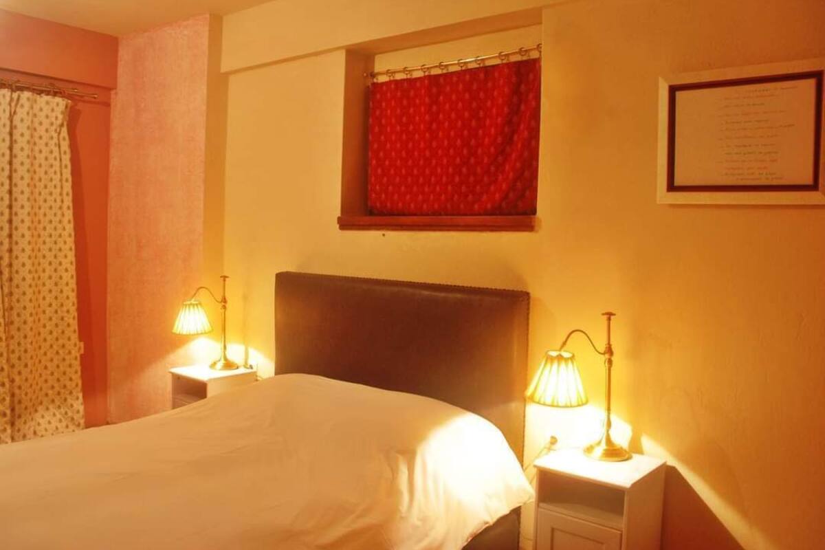 Habitación de hotel con cama, dos lámparas de noche, una ventana con cortina roja y un certificado enmarcado en la pared.