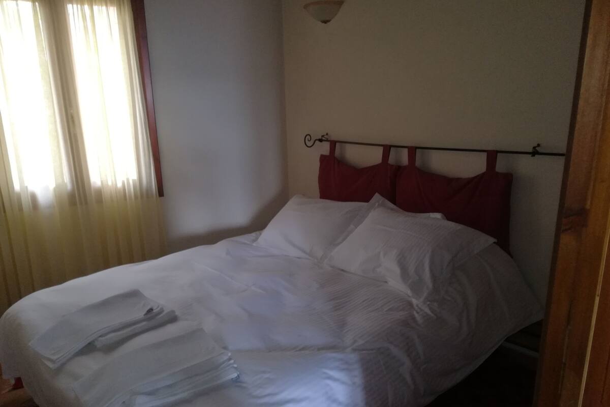 Una cama bien hecha con sábanas blancas y un cabecero rojo, una ventana con cortinas de encaje, y una lámpara fijada a la pared.